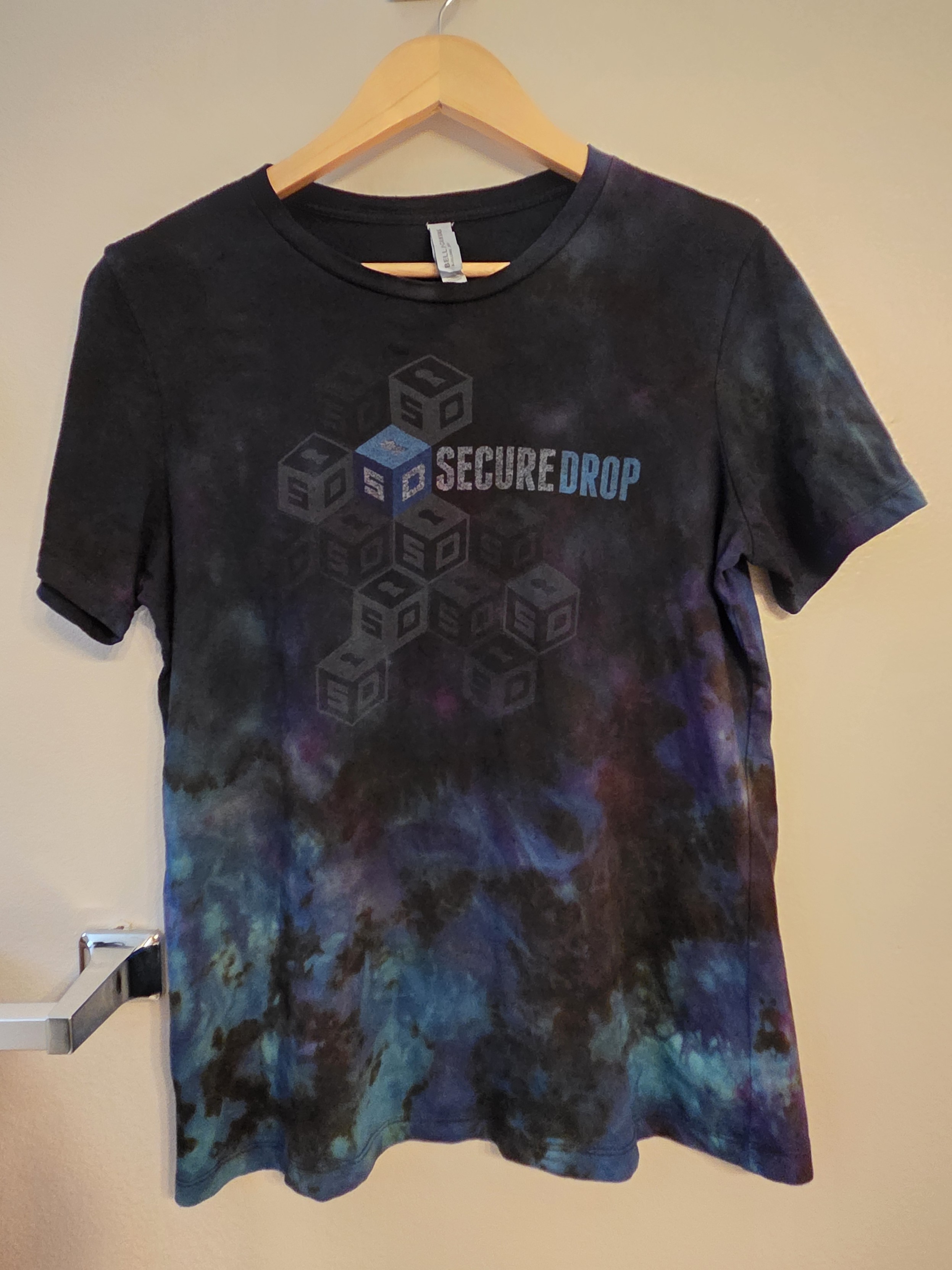 A t-shirt on a hanger, sporting the SecureDrop logo. The shirt itself is no longer the standard issue black, but has been haphazardly lifted and re-dyed a combination of purple, dark blue, and some cyan, hopefully giving the impression that the logo is floating in space with a pretty nebula or spacial anomaly behind and slightly below it.
